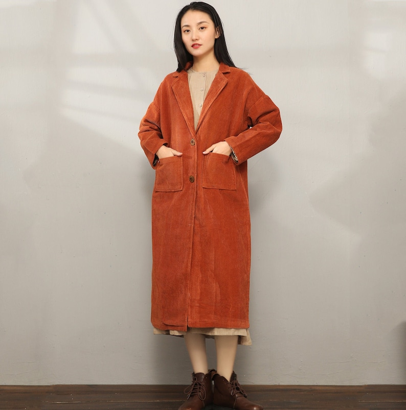 May include: A long, rust-colored corduroy coat with a single button closure and two large patch pockets. The coat has a classic collar and a relaxed fit.