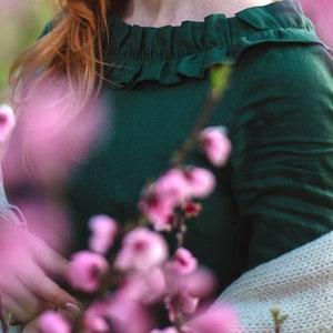 Puede incluir: Una mujer con un vestido verde oscuro con cuello de volantes, de pie frente a un arbusto de flores rosas. Lleva una chaqueta de punto de color crema.
