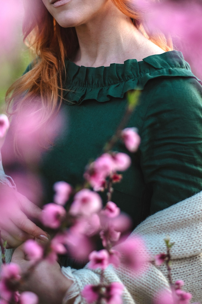 Puede incluir: Una mujer con un vestido verde oscuro con cuello de volantes, de pie frente a un arbusto de flores rosas. Lleva una chaqueta de punto de color crema.