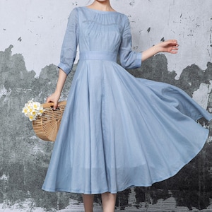 May include: A light blue midi dress with a pleated bodice, three-quarter sleeves, and a flowing skirt. The dress is cinched at the waist and paired with white sandals. A woven basket with flowers is held in the hand.
