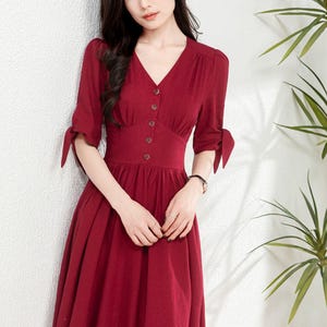 May include: A woman wearing a maroon midi dress with a v-neckline and button closure. The dress has a fitted bodice and a flowy skirt. The sleeves are three-quarter length and have a tie detail at the cuffs.