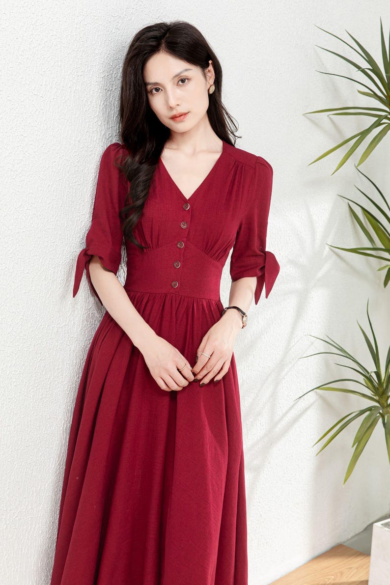 May include: A woman wearing a maroon midi dress with a v-neckline and button closure. The dress has a fitted bodice and a flowy skirt. The sleeves are three-quarter length and have a tie detail at the cuffs.