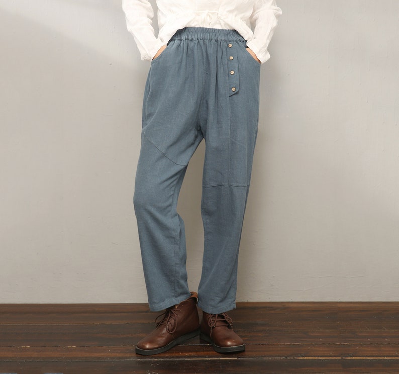 May include: A pair of blue linen pants with a button fly and an elastic waistband. The pants are loose fitting and have a relaxed silhouette. The model is wearing brown leather boots.