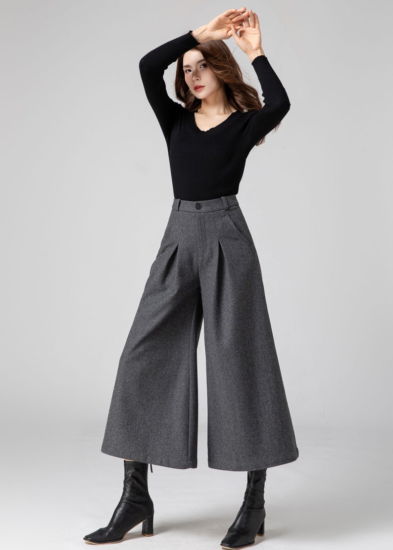 Pu&ograve; includere: Una donna indossa una camicia nera a maniche lunghe e pantaloni larghi grigi. I pantaloni hanno la vita alta e sono plissettati. Indossa stivali neri con tacco.