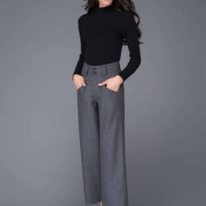 Puede incluir: Una mujer con un jersey negro de cuello alto y pantalones grises de pierna ancha con cintura alta y tres botones. Lleva tacones negros.