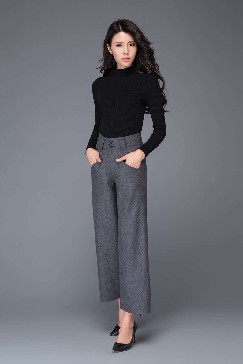 Puede incluir: Una mujer con un jersey negro de cuello alto y pantalones grises de pierna ancha con cintura alta y tres botones. Lleva tacones negros.