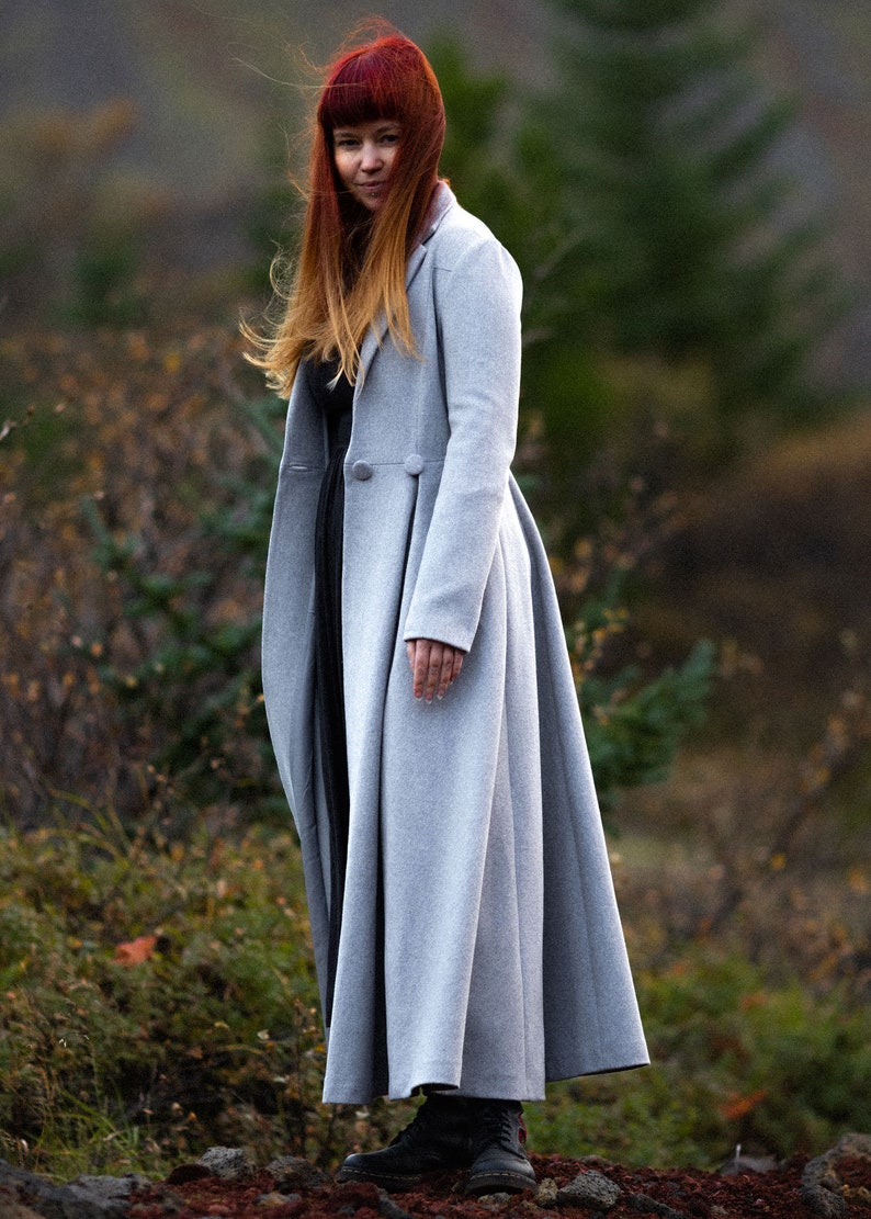 May include: A light gray, long wool coat with a flared skirt and two decorative buttons. The coat is worn over a black dress and paired with black boots. The person has red hair.