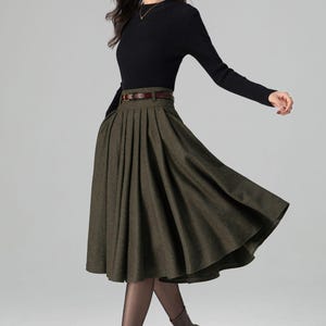 May include: A woman's outfit featuring a black long-sleeve top and a pleated olive green skirt. The skirt is cinched at the waist with a brown belt. The outfit is completed with brown boots.