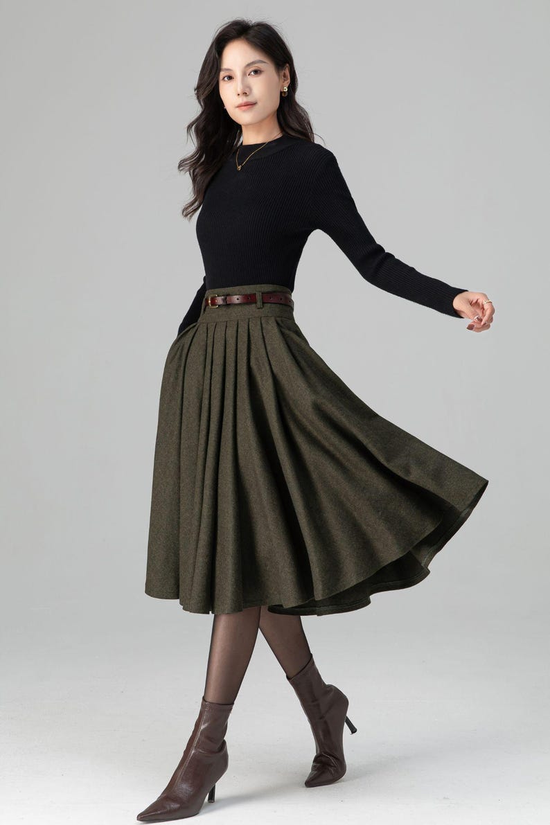 May include: A woman's outfit featuring a black long-sleeve top and a pleated olive green skirt. The skirt is cinched at the waist with a brown belt. The outfit is completed with brown boots.