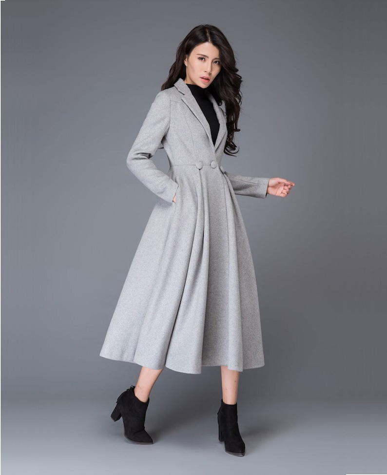 May include: A light gray, knee-length coat with a tailored blazer-style top and a flared skirt. The coat features a two-button closure, long sleeves, and pockets. The model is wearing black ankle boots.