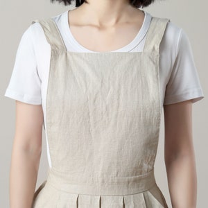 May include: A beige linen overall dress with thin straps. The dress has a pleated skirt and a simple, boxy bodice.