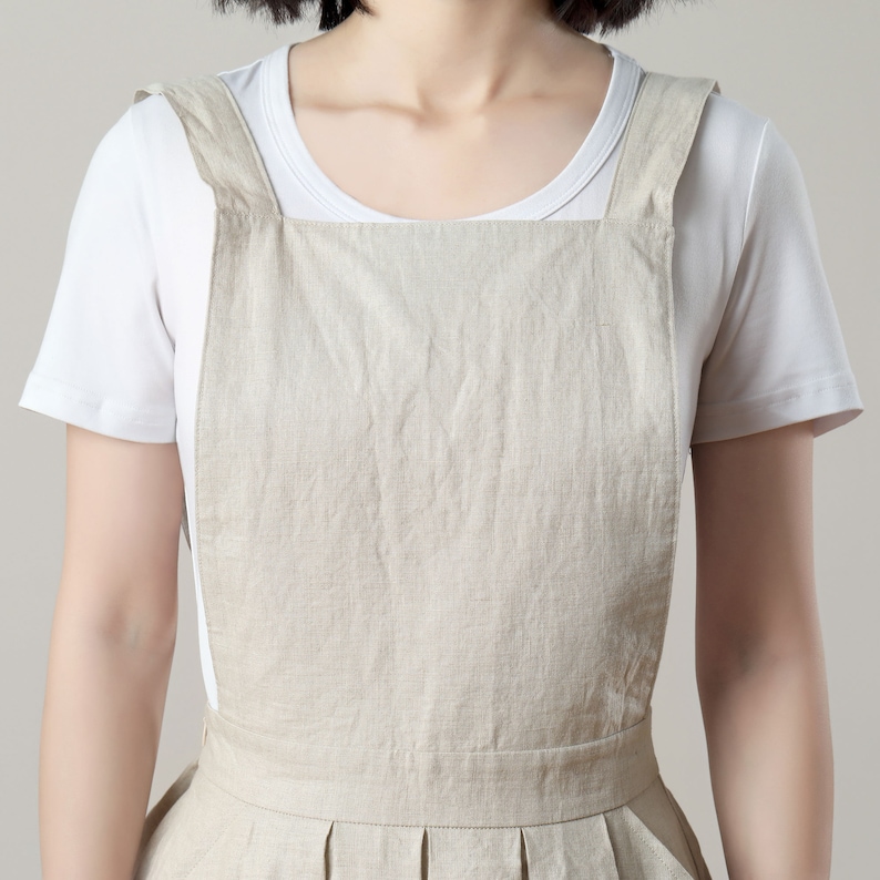May include: A beige linen overall dress with thin straps. The dress has a pleated skirt and a simple, boxy bodice.