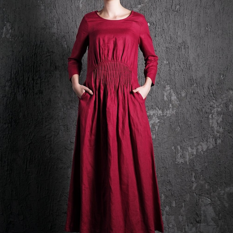 May include: A long, red linen dress with a gathered waist and 3/4 sleeves. The dress has pockets on either side.