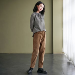 May include: A woman wearing a grey long-sleeved shirt with a lace collar and brown corduroy trousers. She is standing in a room with a green wall and a window in the background.