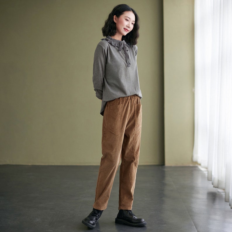 May include: A woman wearing a grey long-sleeved shirt with a lace collar and brown corduroy trousers. She is standing in a room with a green wall and a window in the background.