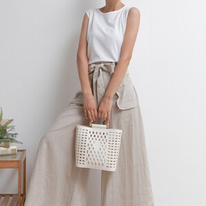May include: A woman wearing a white sleeveless top and beige wide-leg pants with a white woven basket bag with wooden handles.