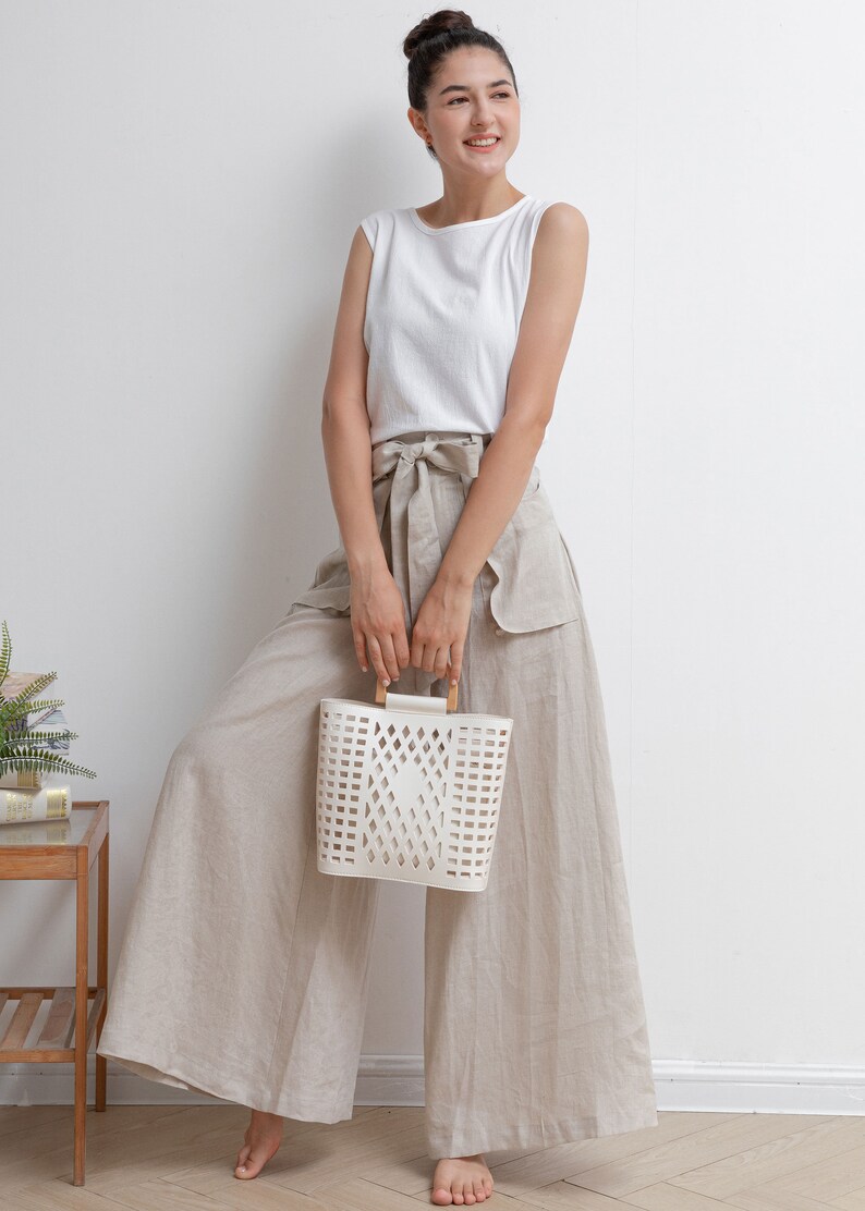 May include: A woman wearing a white sleeveless top and beige wide-leg pants with a white woven basket bag with wooden handles.