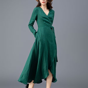May include: A woman wearing a green wrap dress with long sleeves and a tie at the waist. She is wearing a straw hat and black sandals.