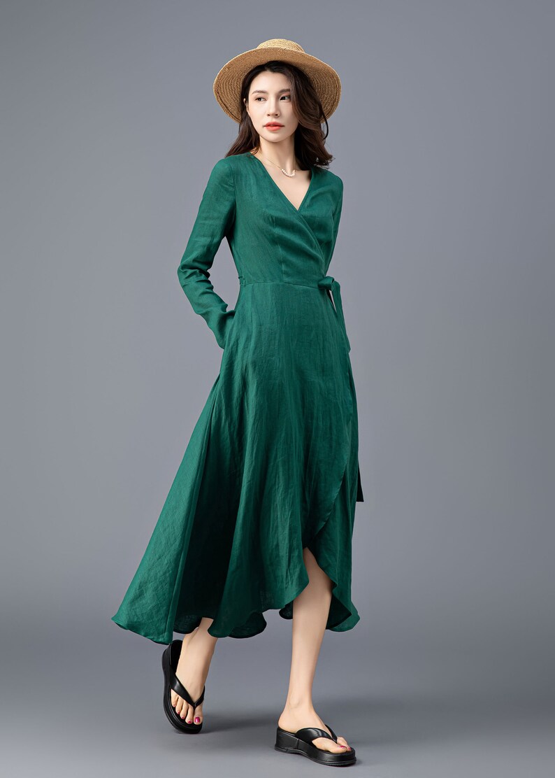 May include: A woman wearing a green wrap dress with long sleeves and a tie at the waist. She is wearing a straw hat and black sandals.