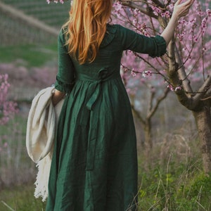Puede incluir: Una mujer con el pelo largo y rojo lleva un vestido verde con una chalina blanca sobre el brazo. Est&aacute; de pie en un campo de flores silvestres.