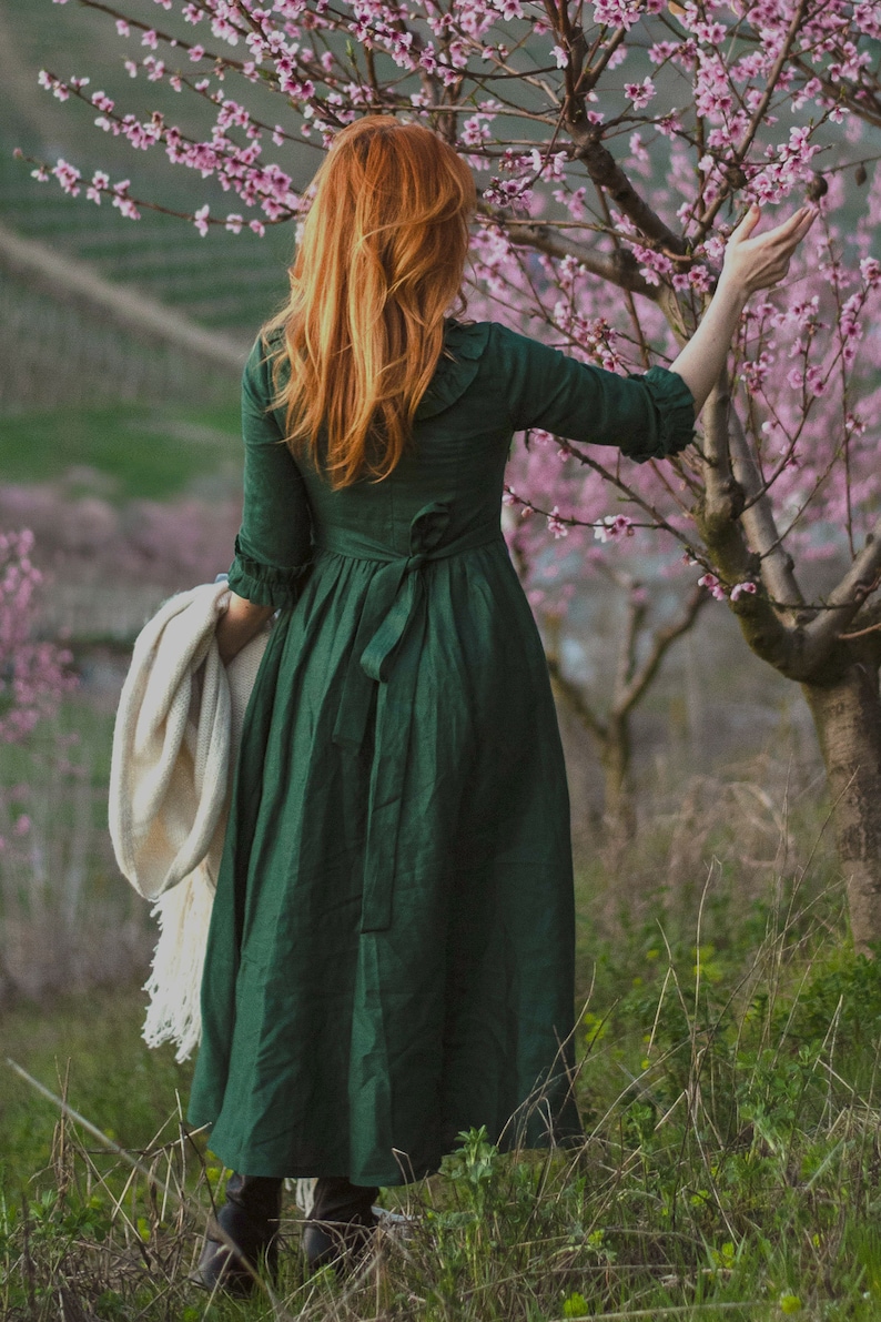 Puede incluir: Una mujer con el pelo largo y rojo lleva un vestido verde con una chalina blanca sobre el brazo. Est&aacute; de pie en un campo de flores silvestres.