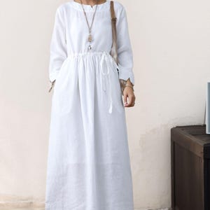 May include: A white linen dress with long sleeves and a drawstring waist. The dress has a relaxed fit and a slightly A-line silhouette. The model is wearing brown leather ankle boots.
