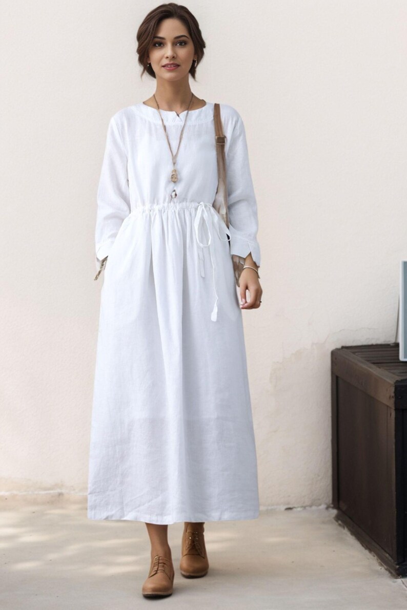 May include: A white linen dress with long sleeves and a drawstring waist. The dress has a relaxed fit and a slightly A-line silhouette. The model is wearing brown leather ankle boots.