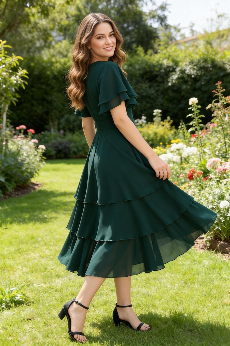 May include: A dark green, tiered midi dress with flutter sleeves. The dress is made of a flowing fabric and is paired with black heeled sandals. The background features a garden with green grass and colorful flowers.