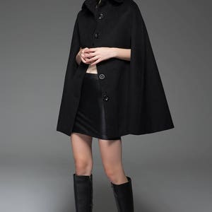 May include: A black wool cape with a collar and button fastening. The cape is worn over a black skirt and black knee-high boots.