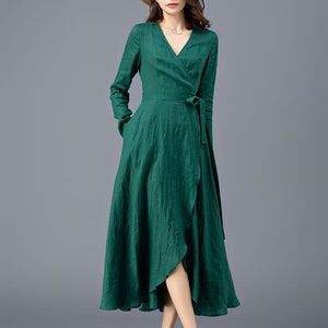 May include: A woman wearing a long-sleeved, emerald green wrap dress with a V-neckline and a matching tie belt. She is wearing a straw hat and black sandals.