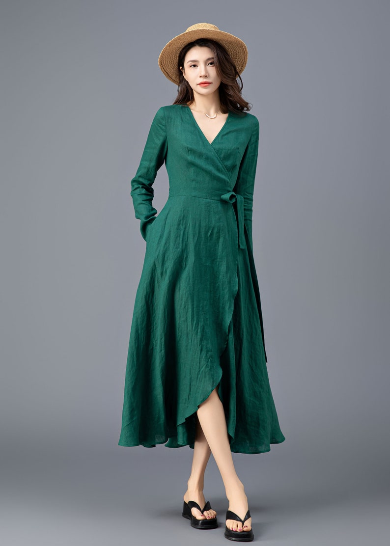 May include: A woman wearing a long-sleeved, emerald green wrap dress with a V-neckline and a matching tie belt. She is wearing a straw hat and black sandals.