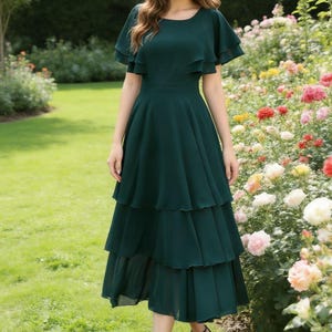 May include: A dark green tiered dress with flutter sleeves. The dress has a fitted waist and a knee-length hemline. The person is wearing black sandals. The background features a garden with green grass and colorful flowers.
