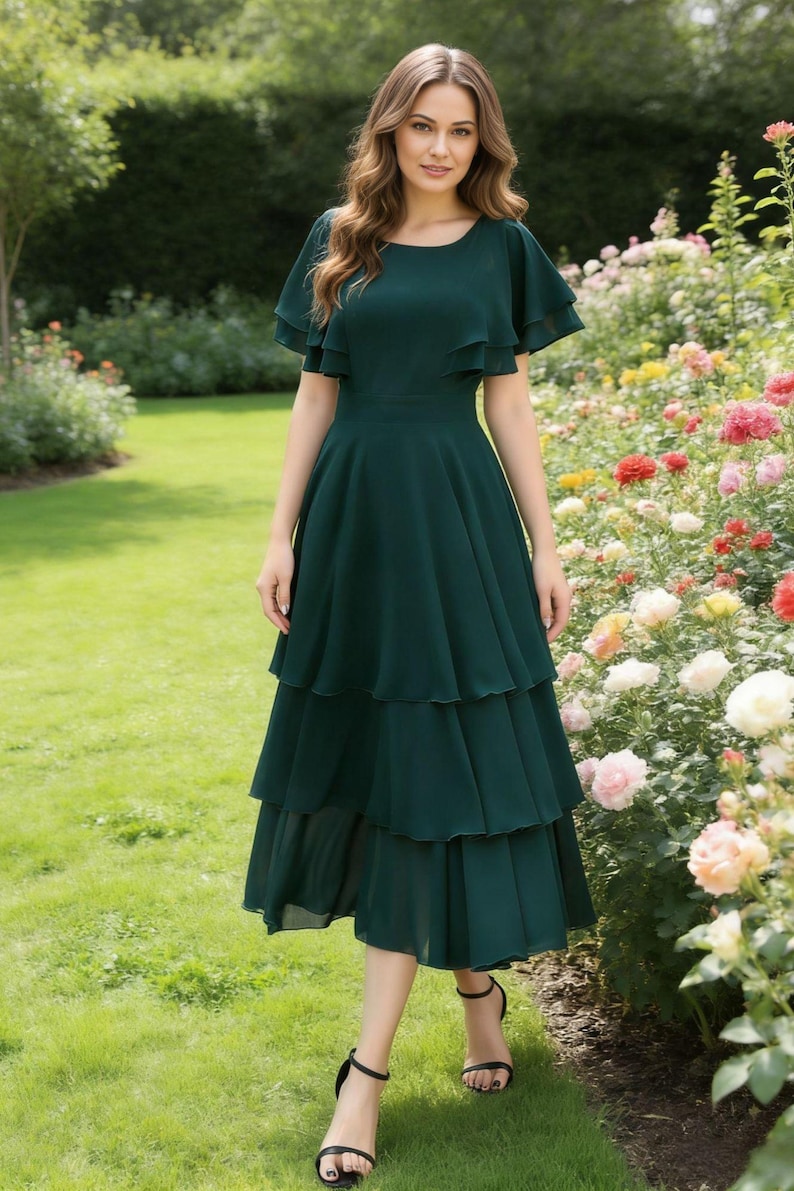 May include: A dark green tiered dress with flutter sleeves. The dress has a fitted waist and a knee-length hemline. The person is wearing black sandals. The background features a garden with green grass and colorful flowers.