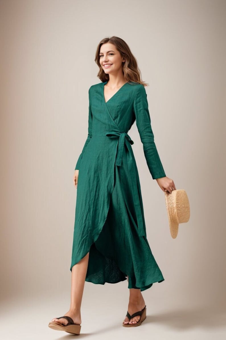 May include: A woman wearing a green wrap dress with long sleeves and a tie at the waist. She is holding a straw hat in her right hand and walking towards the camera.