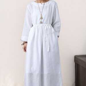 May include: A white linen dress with long sleeves and a drawstring waist. The dress has a relaxed fit and a simple, elegant design.