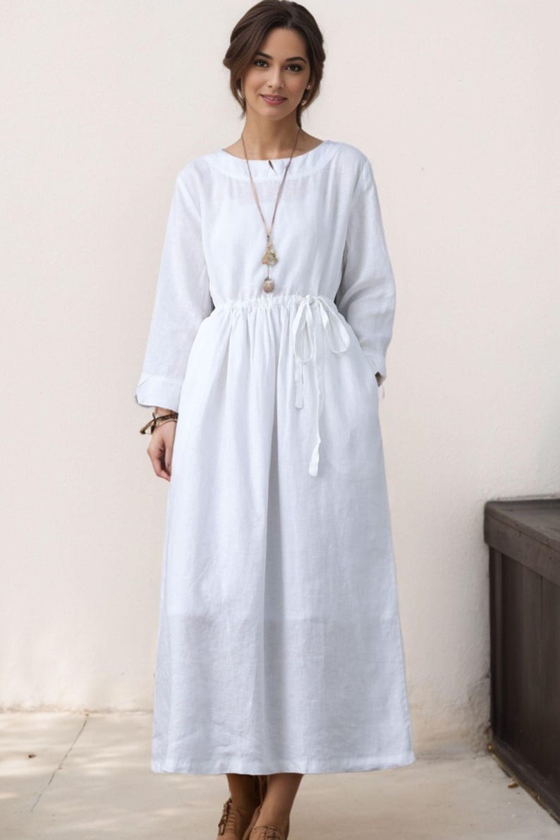 May include: A white linen dress with long sleeves and a drawstring waist. The dress has a relaxed fit and a simple, elegant design.