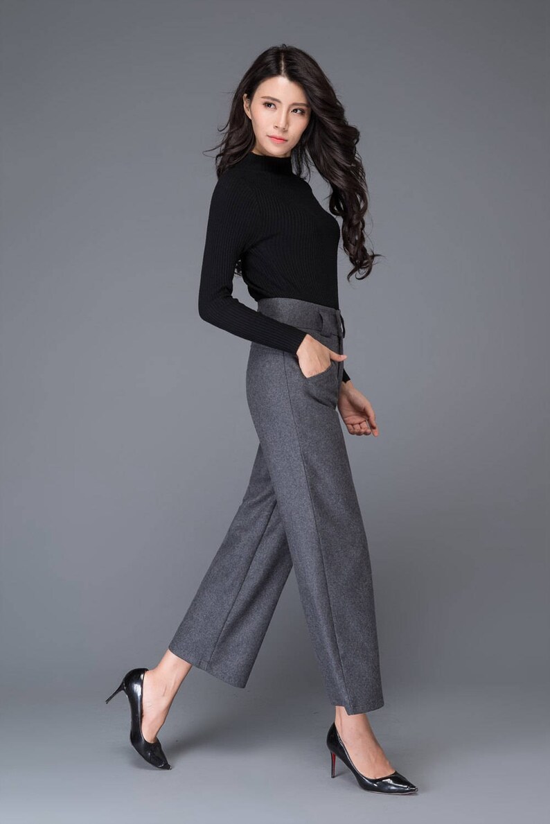Puede incluir: Una mujer con un jersey negro de cuello alto y pantalones grises de pierna ancha con cintur&oacute;n. Lleva tacones altos negros.