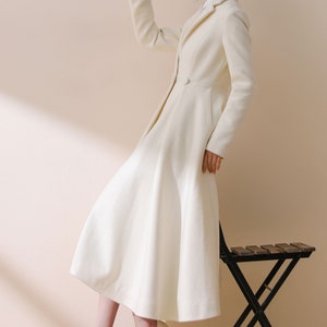 May include: A white long-sleeved coat with a flared skirt and a single button closure. The coat is worn by a person with a hat and white shoes.