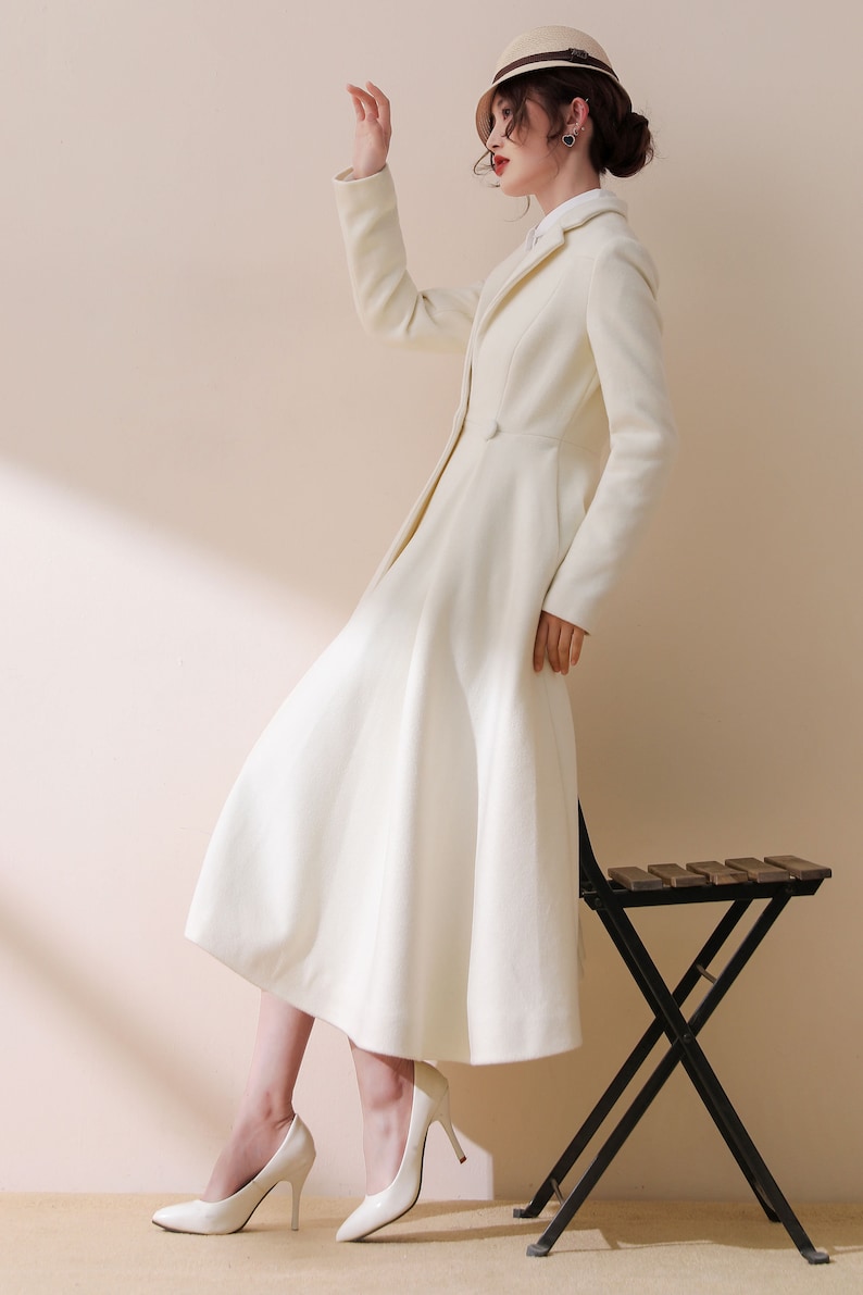 May include: A white long-sleeved coat with a flared skirt and a single button closure. The coat is worn by a person with a hat and white shoes.