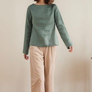 May include: A woman is wearing a sage green linen top with long sleeves and a relaxed fit. She is also wearing beige wide-leg pants.