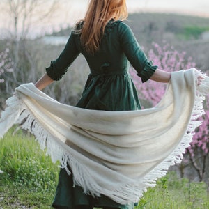 Puede incluir: Una mujer con un vestido verde y una chalina blanca con flecos se encuentra en un campo de hierba verde y flores rosas.