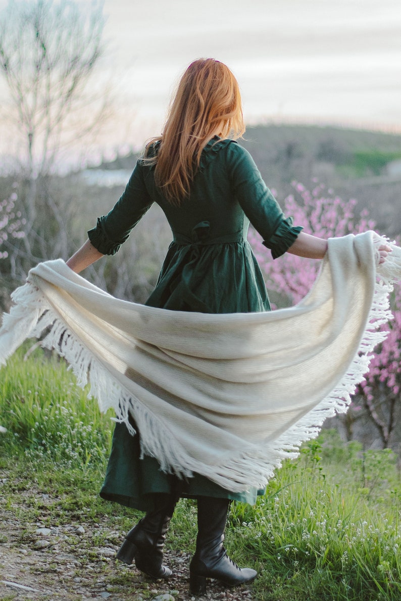 Puede incluir: Una mujer con un vestido verde y una chalina blanca con flecos se encuentra en un campo de hierba verde y flores rosas.