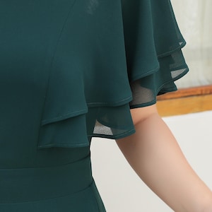 May include: A dark green dress featuring layered, flutter sleeves. The dress has a fitted bodice and a defined waistline. The sleeves are made of a sheer, flowing fabric, adding a touch of elegance. The dress is suitable for formal events.