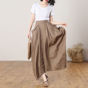 May include: A brown linen maxi skirt with a high waist and pleats. The skirt has a slightly asymmetrical hemline and is made of a lightweight, breathable fabric.