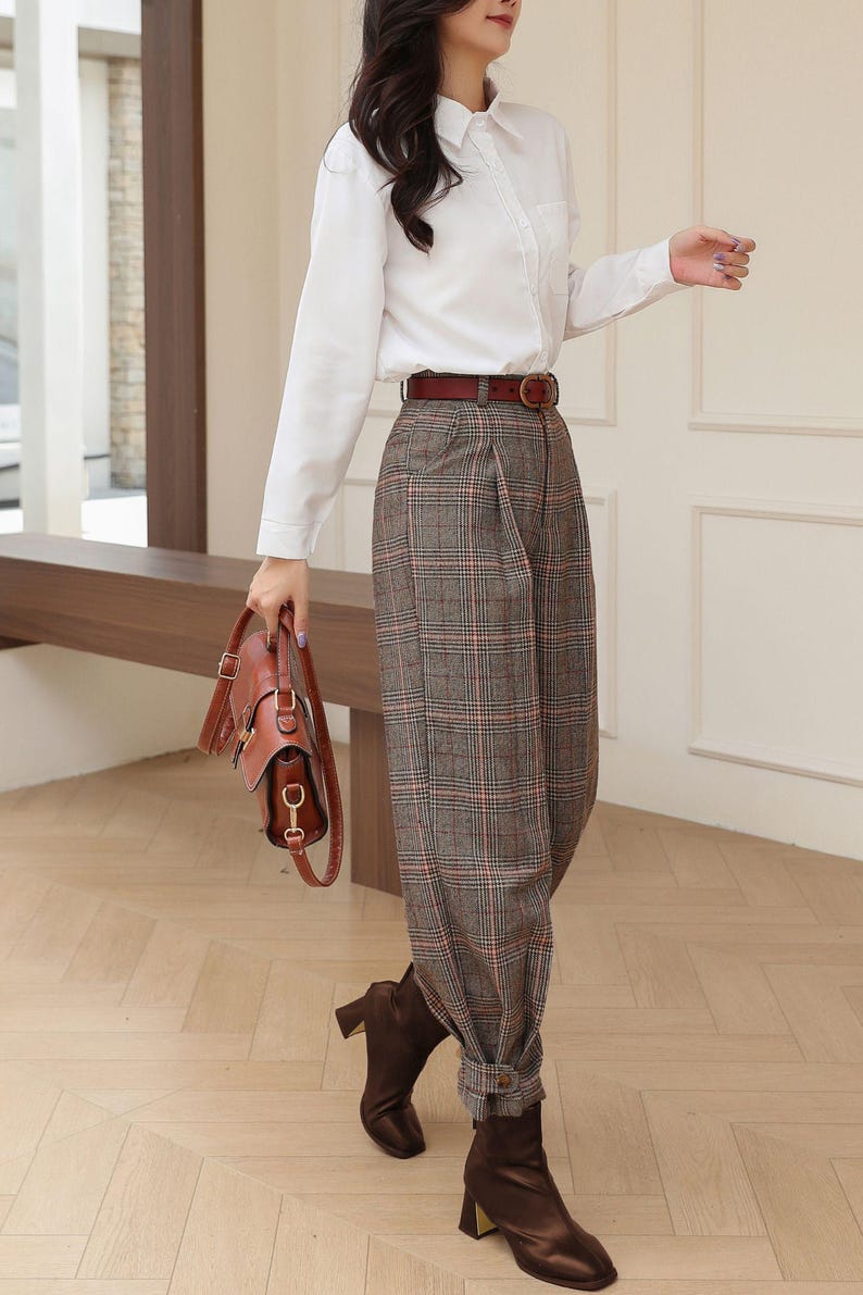 K&ouml;nnte beinhalten: Eine Person tr&auml;gt ein wei&szlig;es Hemd, eine braune karierte Hose und braune Lederstiefel. Ein brauner Lederg&uuml;rtel und eine Handtasche vervollst&auml;ndigen das Outfit. Die Hose hat eine plissierte Vorderseite und ist an den Kn&ouml;cheln umgeschlagen.