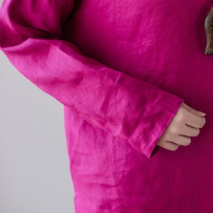 May include: Close-up of a long-sleeved magenta shirt. The fabric appears to be linen, with visible texture and slight wrinkles. The sleeve is visible, with the cuff and part of the hand. The background is a plain, light gray.