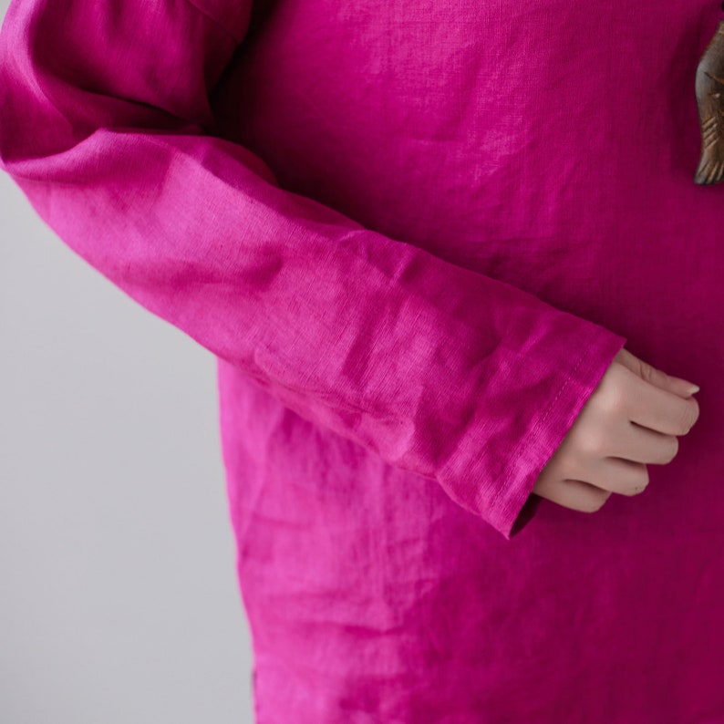 May include: Close-up of a long-sleeved magenta shirt. The fabric appears to be linen, with visible texture and slight wrinkles. The sleeve is visible, with the cuff and part of the hand. The background is a plain, light gray.