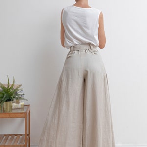 May include: A woman wearing a white sleeveless top and wide-legged, light beige linen pants with a wide belt.