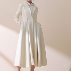 May include: A white long-sleeved coat with a flared skirt and a button closure. The coat has a classic collar and two pockets. The model is wearing a white shirt and a straw hat with a brown ribbon.