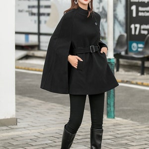 May include: A black wool cape with a high collar, a belt, and a button closure. The cape is worn over a black mock neck shirt and black pants. The person is wearing brown boots and a brown newsboy cap.